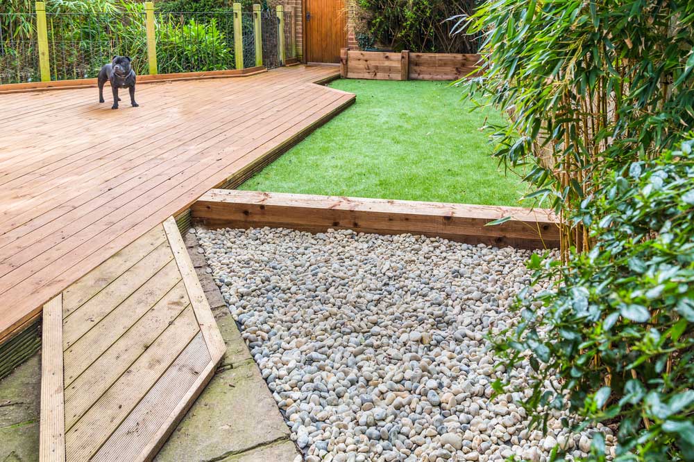 A section of a residential garden, yard with wooden decking, patio over a fish pond, a section of artificial grass and an area of stone pebble. There is a bamboo plant and a dog in the garden.